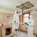 Workers in protective gear assessing asbestos risk in a room with damaged ceiling and caution signs