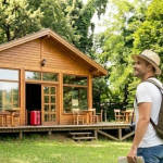 Man with backpack and hat stands outside a wooden cabin surrounded by lush greenery